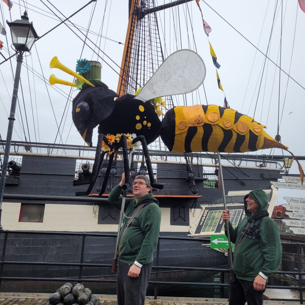 Photo of two puppeteers carrying a large puppet wasp insect above their heads on poles in front of a ship