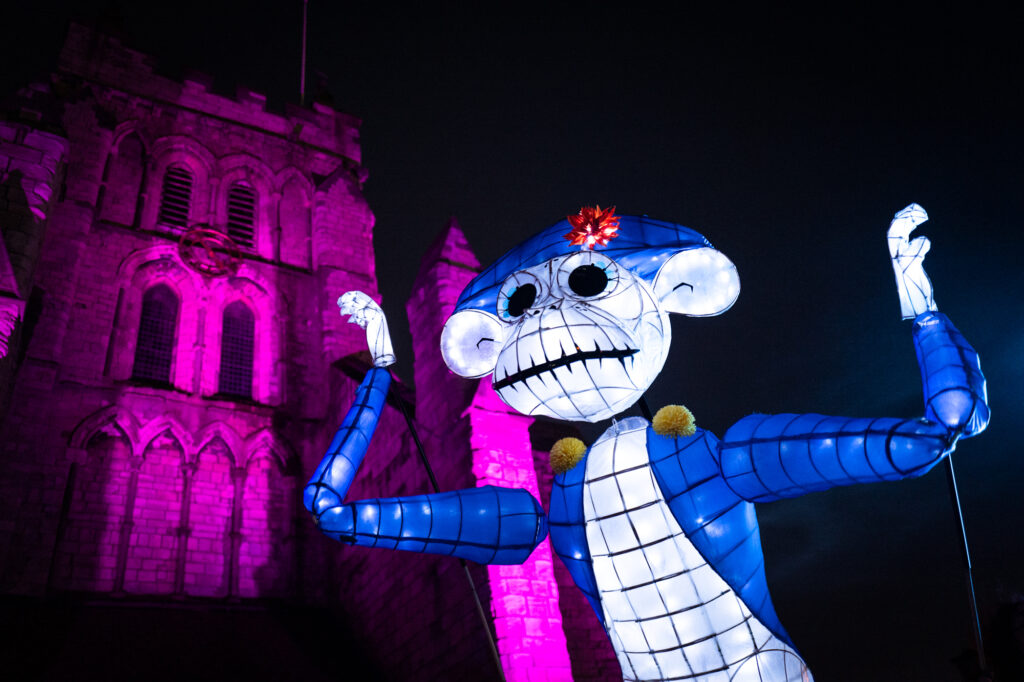 Large scale monkey lantern puppet wearing blue tail coat and bicorn hat illuminated at night against a pink lit church