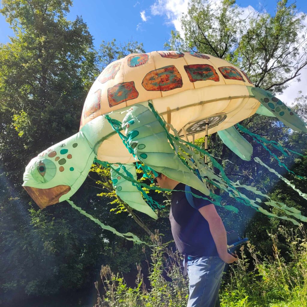 Photo of a large inflatable sea turtle carried on a backpack in a park. Trees in the background on a blue sky sunny day.