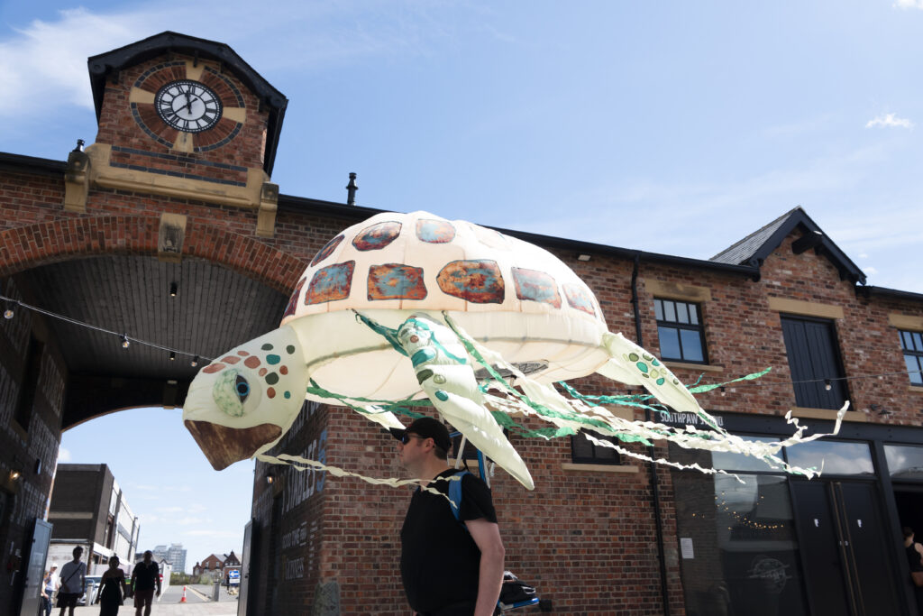 An inflatable sea turtle animal carried on a backpack above the head, outdoor at a festival
