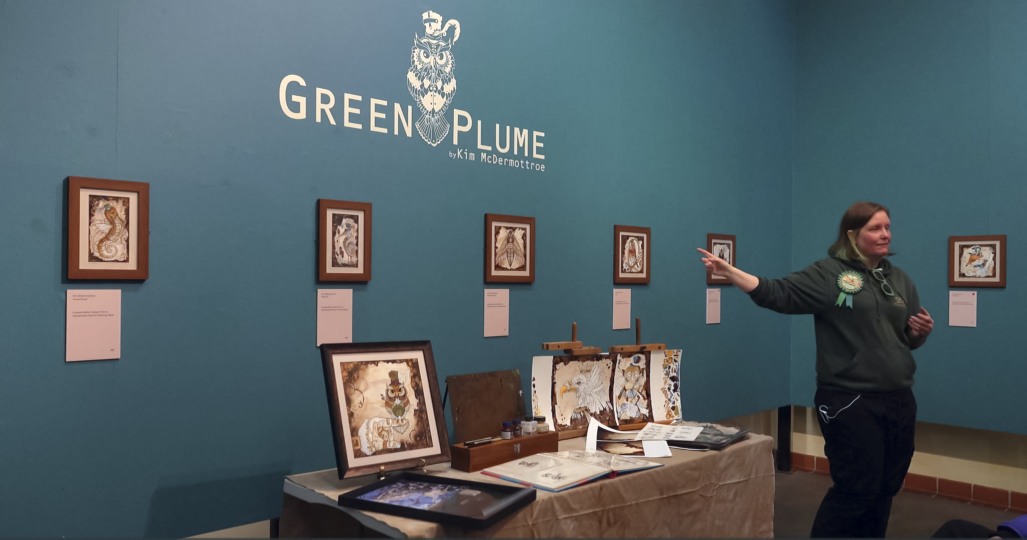 Kim is standing in front of her illustrations at her exhibition in an art gallery. She's doing an artist talk and pointing to the framed illustrations on the wall.